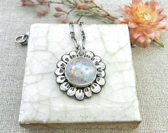 May include: A silver pendant necklace with a round, iridescent glass center. The pendant is surrounded by a detailed, scalloped silver frame. The necklace rests on a white, cracked tile surface, with a flower and sprig in the background.