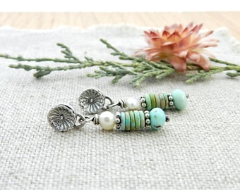 May include: A pair of dangle earrings featuring silver-tone floral studs, white pearls, and turquoise and green beads. The earrings are displayed on a textured, light-colored surface with a sprig of greenery and a pink flower in the background.