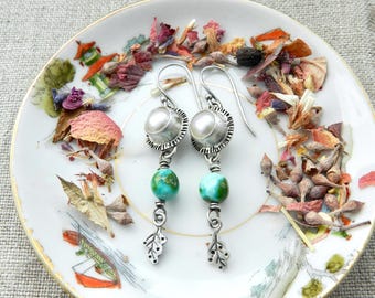 May include: A pair of silver earrings with a pearl and turquoise bead design. The earrings are on a white plate with floral and leaf accents. The plate is surrounded by dried flowers and leaves.