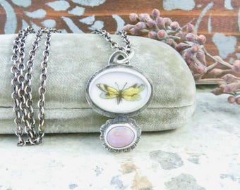 May include: A silver-toned pendant necklace featuring an oval-shaped charm with a butterfly illustration in yellow and green. Below the charm is a round, light pink stone. The necklace chain is a dark silver color.