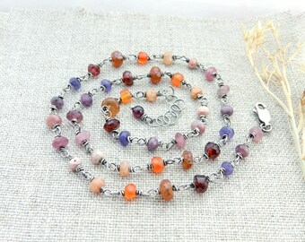 May include: A colorful necklace featuring a variety of gemstone beads in shades of pink, purple, orange, and red. The beads are strung on a silver-colored chain and secured with a clasp. The necklace is displayed on a textured, light-colored surface.