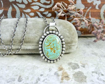 May include: Oval turquoise pendant necklace with a silver chain. The pendant features a light blue stone with brown veining, surrounded by a beaded silver frame. The necklace is displayed on a gray velvet surface.