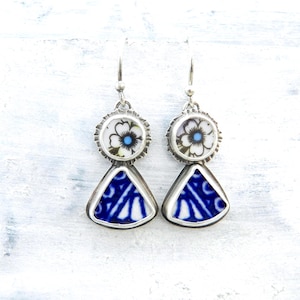 May include: A pair of handcrafted earrings featuring a floral design and blue and white geometric patterns. The earrings have a silver-tone metal setting and a hook closure. The top portion has a white flower with a blue center. The bottom portion is a blue and white triangle.