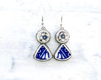 May include: A pair of handcrafted earrings featuring a floral design and blue and white geometric patterns. The earrings have a silver-tone metal setting and a hook closure. The top portion has a white flower with a blue center. The bottom portion is a blue and white triangle.