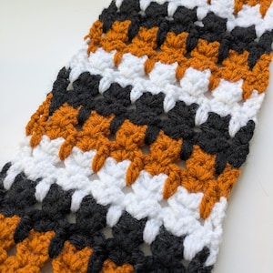 Puede incluir: Una bufanda de ganchillo hecha a mano con un patrón repetido de hilo negro, blanco y naranja. La bufanda tiene un punto texturizado, creando un accesorio visualmente interesante y acogedor. La combinación de colores es otoñal.