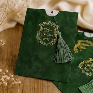 Puede incluir: Bolsa de regalo verde oscuro con diseño dorado y borla. La bolsa presenta el texto "Roberta Christmas 2023" en un marco ornamentado. La borla es del mismo color que la bolsa.