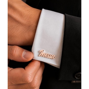 May include: A rose gold-toned metal cufflink featuring the name "Messa" in a cursive script font, attached to the white cuff of a long-sleeved shirt worn under a black jacket. The accessory is secured through the buttonholes of the fabric cuff.