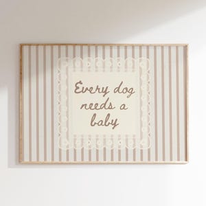 Op de afbeelding: Een ingelijste print met een lichtbruine houten lijst. De print heeft een crèmekleurige achtergrond met verticale strepen in lichtbruin en wit. De tekst "Every dog needs a baby" staat in een decoratieve rand.