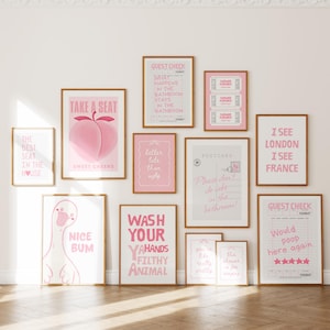 Könnte beinhalten: Eine Galeriewand mit elf gerahmten Drucken in hellen Holzrahmen, die rosa-weiße Designs mit humorvollen Texten und Illustrationen wie einen Pfirsich, eine Gans, Gästeschecks und Postkarten auf einer schlichten Wand zeigen.