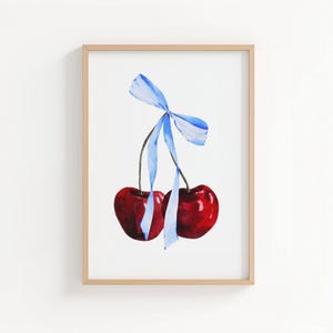 Peut inclure: Une impression aquarelle encadrée représentant deux cerises rouges brillantes reliées par leurs tiges, nouées ensemble avec un ruban bleu clair. L'œuvre est présentée dans un cadre en bois clair simple sur un fond blanc uni.