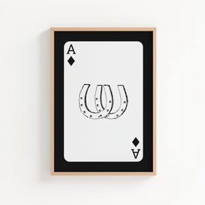May include: A framed print of the Ace of Diamonds playing card. The card features a white background with a black border and a black diamond at the top and bottom. Three black horseshoes are in the center of the card.