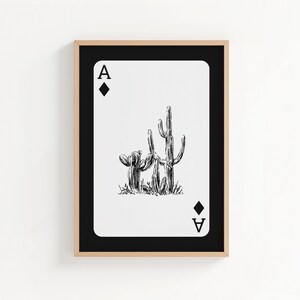 May include: Framed art print featuring a playing card design. The Ace of Diamonds card displays a black and white illustration of two cacti. The card is set against a white background, framed in black and a natural wood tone.