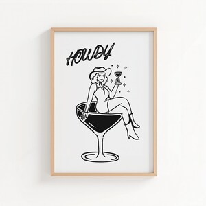 May include: Black and white illustration of a woman in a cowboy hat sitting in a large cocktail glass, holding a drink. The word "Howdy" is written above the woman in a cursive font. The artwork is framed in a light wood frame.