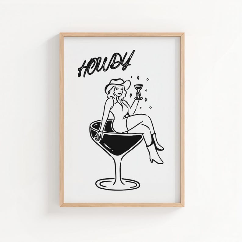 May include: Black and white illustration of a woman in a cowboy hat sitting in a large cocktail glass, holding a drink. The word "Howdy" is written above the woman in a cursive font. The artwork is framed in a light wood frame.