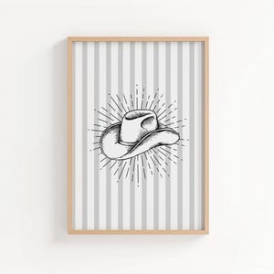 May include: A framed print featuring a black and white illustration of a cowboy hat. The hat is centered on a background of vertical gray and white stripes. The hat is surrounded by a sunburst design.