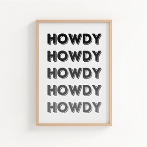 May include: A framed print with the word "HOWDY" repeated five times in a bold, black font. The text is centered on a white background, and the frame is a light wood color. The artwork is a simple, yet eye-catching piece.