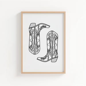 May include: A framed print featuring a pair of cowboy boots. The boots are depicted in a black and white line drawing style, with intricate decorative patterns. The boots are set against a white background, and the frame is a light wood color.
