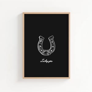 May include: A framed print featuring a white horseshoe illustration on a black background. The words "Lucky you" are written in cursive below the horseshoe. The frame is a light brown color.