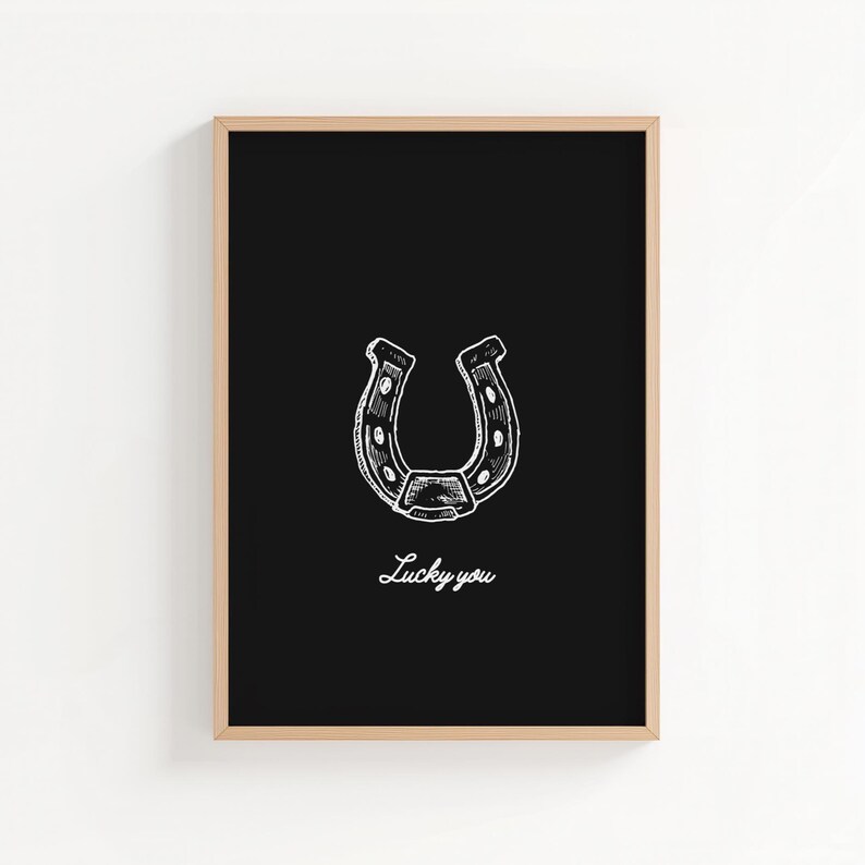 May include: A framed print featuring a white horseshoe illustration on a black background. The words "Lucky you" are written in cursive below the horseshoe. The frame is a light brown color.
