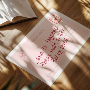 Puede incluir: Una postal rectangular con fondo rosa claro que presenta el texto "...and so together they built a life they loved." La tarjeta incluye un gr&aacute;fico de sello con un coraz&oacute;n, la palabra "POSTCARD" y una l&iacute;nea "FROM:" en tinta roja oscura.