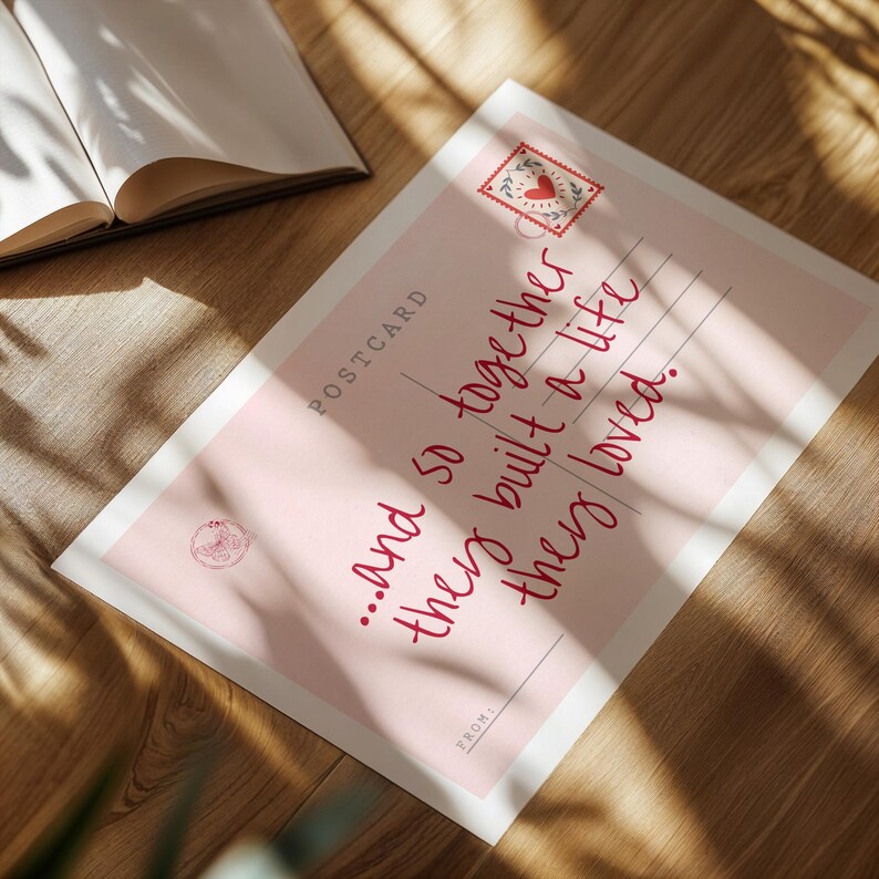 Puede incluir: Una postal rectangular con fondo rosa claro que presenta el texto "...and so together they built a life they loved." La tarjeta incluye un gr&aacute;fico de sello con un coraz&oacute;n, la palabra "POSTCARD" y una l&iacute;nea "FROM:" en tinta roja oscura.