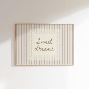 May include: A rectangular wall art print featuring a light beige and white vertical striped background. In the centre, a cream-coloured square with a decorative lace-style border contains the brown handwritten text Sweet dreams.