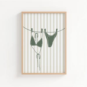 Peut inclure: Une affiche encadrée représentant un ensemble de bikini vert suspendu à une corde à linge. L'illustration est placée sur un fond de rayures verticales beiges et blanches, avec un cadre en bois clair.