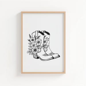 May include: Black and white illustration of two cowboy boots adorned with floral arrangements. The boots are side-by-side, with detailed linework depicting the boot's shape and the flowers' petals and leaves. The artwork is framed in a light wood frame.