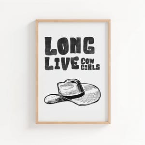 May include: A framed print with the text "LONG LIVE COW GIRLS" in bold, black letters. Below the text is a black and white illustration of a cowboy hat. The frame is a light brown color.