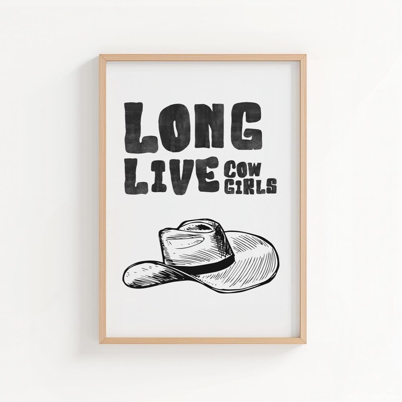 May include: A framed print with the text "LONG LIVE COW GIRLS" in bold, black letters. Below the text is a black and white illustration of a cowboy hat. The frame is a light brown color.
