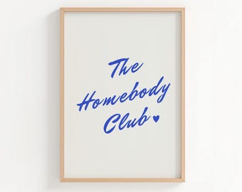 May include: A rectangular wall art print in a light wood frame featuring the blue text The Homebody Club with a small blue heart symbol centred on a plain white background. The design uses a casual, handwritten script font style for the lettering.
