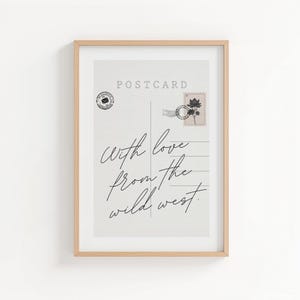 May include: Framed print of a vintage postcard design. The postcard features the word "POSTCARD" at the top, a stamp, and the handwritten phrase "With love from the wild west." The frame is a light brown color.