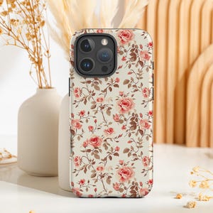 May include: A floral phone case featuring a pattern of pink roses and brown leaves on a cream background. The case has a camera cutout and is designed to fit a smartphone. The case is displayed on a white surface.