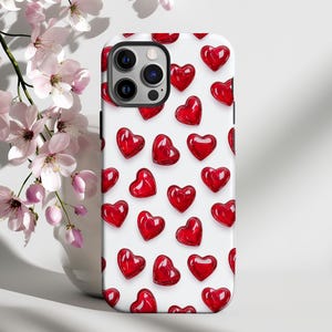 Może przedstawiać: Białe etui na iPhone z wzorem czerwonych serc. Etui ma wycięcie na obiektyw aparatu.