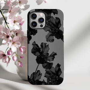 以下が含まれることがあります： 黒い花柄のグレーのiPhoneケース。花はスタイリッシュで、ほぼ黒に近い暗い色をしています。