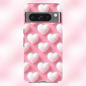 May include: A pink phone case with a pattern of white fuzzy hearts. The case has a black camera lens cutout.