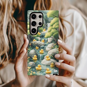 Puede incluir: Una funda para teléfono con una ilustración colorida de un arroyo con patos de goma amarillos flotando en él. La funda tiene un recorte para la cámara y un fondo verde.