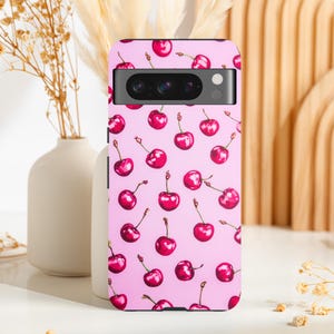 Peut inclure: Une coque de téléphone rose avec un motif de cerises rouges. La coque a une découpe pour l'objectif de l'appareil photo noir.