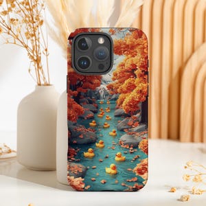 Puede incluir: Una funda para teléfono con una escena colorida de bosque de otoño y un arroyo de patos de goma amarillos. La funda tiene un recorte de cámara negro y un fondo blanco.