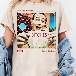 May include: Beige t-shirt with a graphic of Pee-wee Herman holding a sign that says "BITCHES." The shirt also includes the text "1952" and "Paul Reubens."