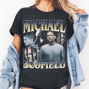 May include: Black t-shirt featuring the name "MICHAEL SCOFIELD" in a gold and purple font. The shirt has a vintage design with multiple images of a man's face. The shirt is worn by a person wearing a denim jacket.