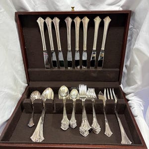 May include: A set of silver-toned flatware arranged in a dark brown wooden storage case with a brown velvet interior. The collection includes eight upright knives and a variety of spoons and forks with fluted handles resting in individual slots.