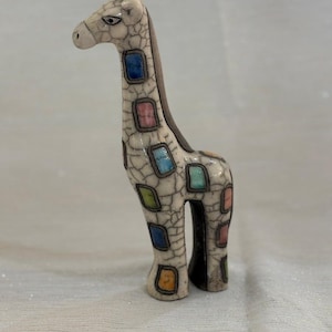 May include: A ceramic giraffe figurine with a cream-colored body and a cracked glaze effect. The giraffe has a dark brown spine and colorful square patches in shades of blue, pink, green, and orange. The figurine is standing upright.