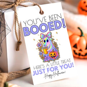 May include: A Halloween gift tag with the text "YOU'VE BEEN BOOED!" and a colorful ghost illustration holding a drink. The tag also says "HERE'S A LITTLE TREAT JUST FOR YOU! Happy Halloween!" and is tied to a white gift box.