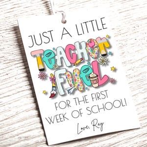 May include: A white gift tag with the text "JUST A LITTLE TEACHER FUEL FOR THE FIRST WEEK OF SCHOOL!" in colorful letters and illustrations. Includes a coffee cup, pencil, and paint brush.
