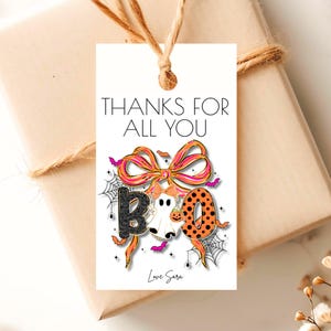May include: A Halloween-themed gift tag with the words "THANKS FOR ALL YOU" at the top. The tag features a colorful "BOO" design with a ghost, spiderwebs, and bats. The tag is tied to a wrapped gift with twine.