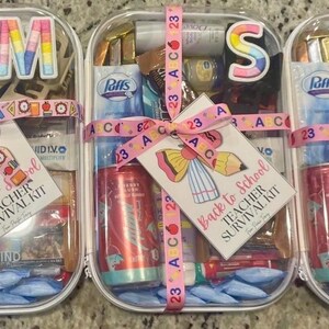 May include: Three clear plastic containers, each a 'Back to School Teacher Survival Kit', filled with items like a red soda can, snacks, and tissues. The kits are tied with pink ribbons and tags. The kits are designed to help teachers.
