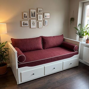Peut inclure: Un lit de repos blanc avec trois tiroirs de rangement, doté de coussins et de traversins rouge foncé avec un passepoil blanc. Le cadre est en bois blanc avec un design à panneaux sur les côtés. Les coussins sont de forme rectangulaire et cylindrique.
