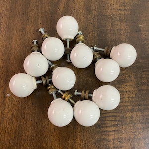 May include: A collection of eleven white ceramic ball cabinet knobs with brass and silver hardware. The knobs are arranged on a dark wood surface, showcasing their round shape and vintage style. Ideal for furniture restoration or adding a classic touch.