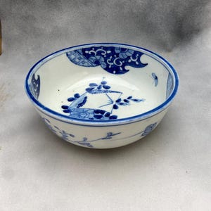 May include: A small, round, white ceramic bowl with a blue rim and floral designs. The bowl features intricate blue patterns, including stylized flowers and cloud-like motifs. The interior has a delicate floral design.